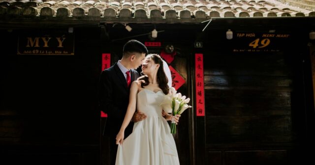 A groom and bride sticking with each other while bride is holding a bouquet of white flowers, with a background of a chinese style house