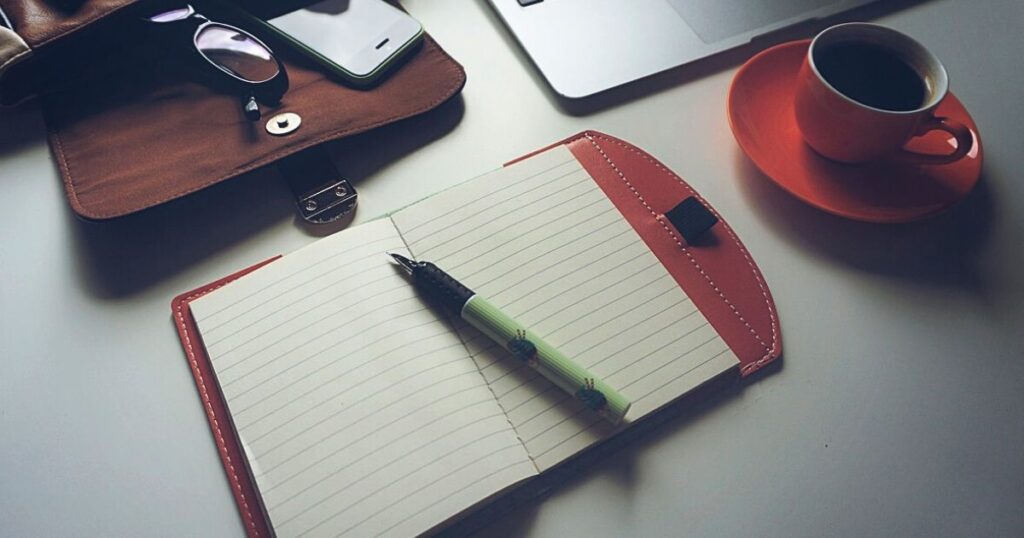 A notebook and a pen on a desk, with a cup of coffee, laptop and a bag of phone and glasses beside the notebook